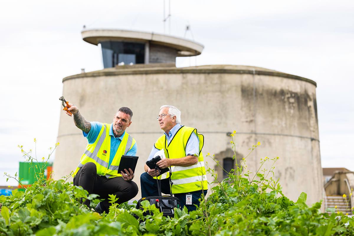 Two male Mitie engineers wearing high vis vests, crouching in front of Martello Tower in Essex. 右边的人手里拿着工具指着屏幕外